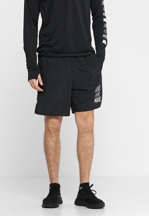 Mann trägt schwarzes Langarmshirt und schwarze Nike-Shorts mit Logo, steht mit Händen in den Taschen und schwarzen Sportschuhen vor weißem Hintergrund.