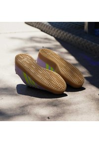 Płaskie buty wsuwane z fioletową cholewką, ozdobione zielonymi paskami. Gruba, teksturowana gumowa podeszwa z falistym wzorem zapewniającym przyczepność.