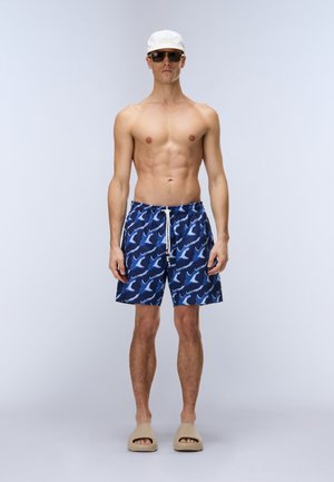 Mężczyzna bez koszulki, w białej czapce, ciemnych okularach przeciwsłonecznych, niebieskich kąpielówkach z wzorem rekina oraz beżowych klapkach stojący na jednolitym tle.