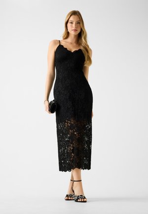 Schwarzes Spitzenkleid mit einem geschwungenen Ausschnitt, tailliertem Oberteil und durchsichtigen Spitzenakzenten am Saum. Kombiniert mit schwarzen, verzierten Sandalen.