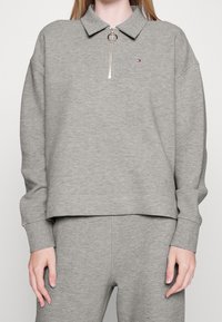 Suéter gris con cuello tipo polo y cremallera. Textura acanalada, corte holgado y un pequeño logo en el pecho. Mangas largas y dobladillo relajado.