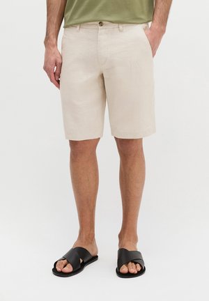 Mann trägt beigefarbene knielange Shorts mit Fronttaschen, ein grünes Hemd, das hineingesteckt ist, und schwarze offene Sandalen mit Zehenfrei vor einem einfarbigen Hintergrund.