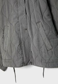 Veste matelassée grise avec piqûres en forme de vagues, deux poches frontales verticales, boutons-pression et cordons de serrage à l'ourlet sur fond blanc.