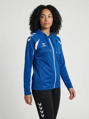 Kobieta o kręconych włosach ubrana w niebieską sportową bluzę z zamkiem i czarne spodnie z białym wzorem w jodełkę, stojąca na jednolitym tle.