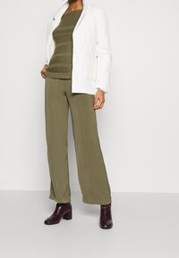 Gesteppte weiße Jacke, olivgrüner gestreifter Pullover, weite olivgrüne Hose und bordeauxrote Stiefel mit Blockabsatz. Durchgehend glatte Texturen.