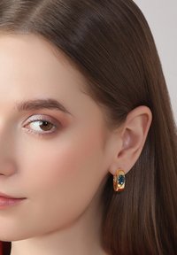 Goldfarbene Creolen mit einem tealfarbenen Edelsteinakzent, glatter Metalloberfläche und kreisförmigem Design, die vor glattem braunen Haar zur Geltung kommen.