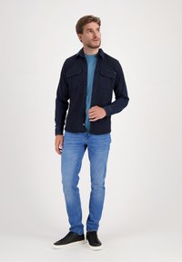 Camisa azul marino de botones con dos bolsillos en el pecho, usada sobre una camiseta azul verdosa, combinada con jeans de un azul claro y zapatillas negras. Textura suave.