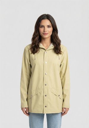 Donna che indossa un impermeabile con cappuccio di colore beige chiaro, con bottoni a pressione e tasche con pattina frontali, in piedi davanti a uno sfondo grigio uniforme.