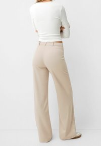 Beige vida byxor med slät tyg, hög midja och en klassisk design. Bakfickor ingår, kombinerat med en åtsittande vit topp.