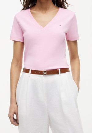 Femme portant un t-shirt rose clair à col en V manches courtes avec un petit logo et un pantalon blanc taille haute avec une ceinture marron.
