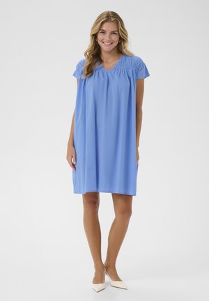 Smilende kvinde står iført en løs, kortærmet, lyseblå kjole og hvide, spidse sko mod en ensfarvet lys baggrund.