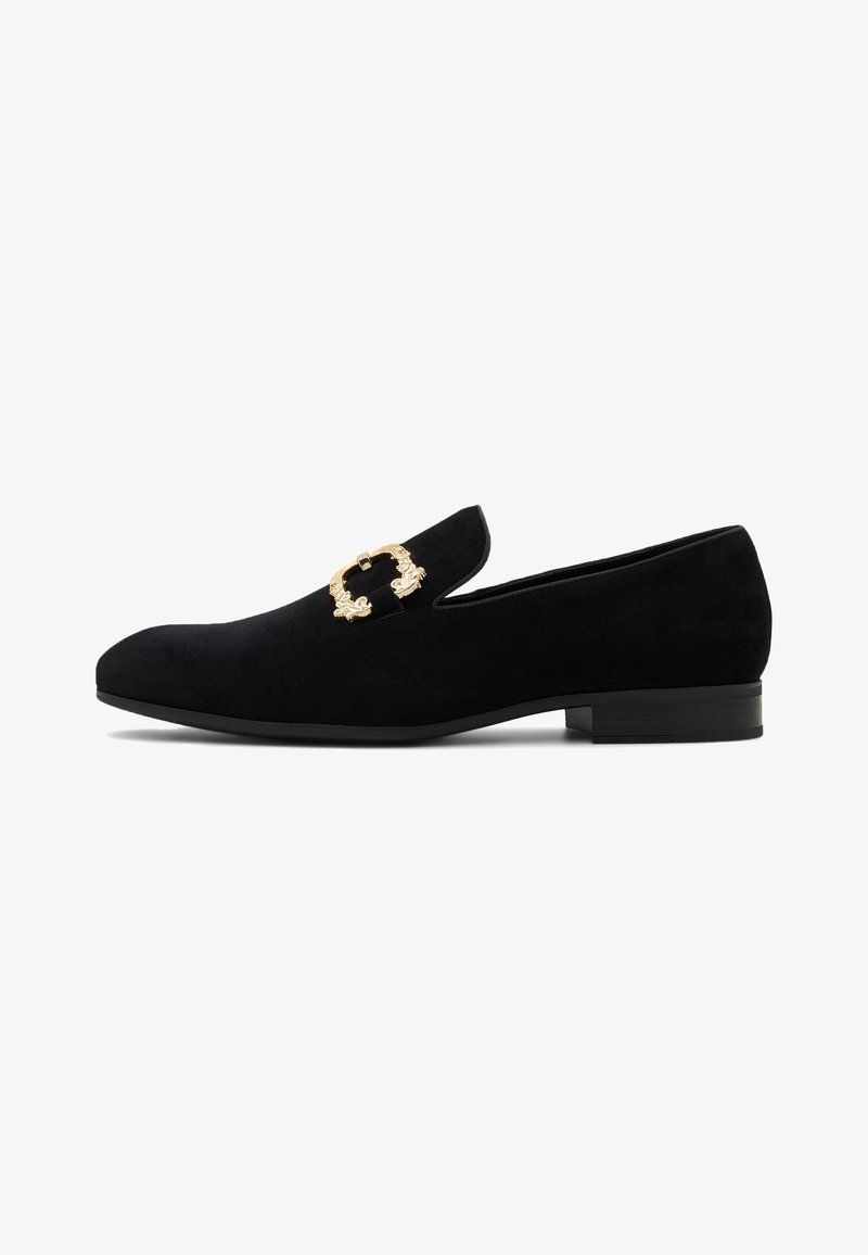 Mocassin homme en suède noir avec un petit talon et un détail décoratif en boucle dorée sur le dessus, sur fond blanc.