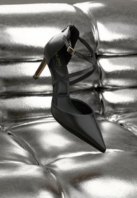 Svart spisshøyhælte sko med kryssede stropper og gullspenne som hviler på en blank sølvfarget, tuftet overflate.