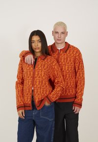 Cardigans zippés en tricot de couleur orange vif avec des motifs géométriques, des poignets côtelés et des accents au niveau du col. Associés à un pantalon bleu et noir.