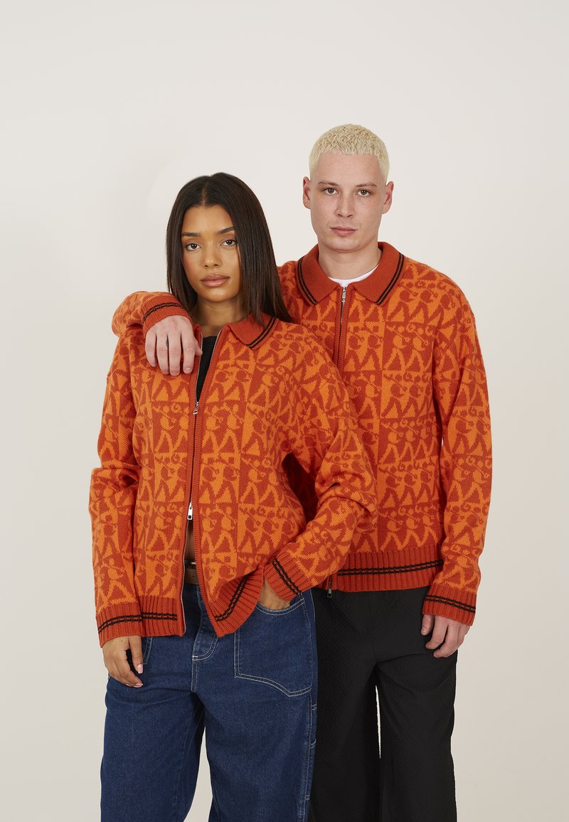 Cardigans zippés en tricot de couleur orange vif avec des motifs géométriques, des poignets côtelés et des accents au niveau du col. Associés à un pantalon bleu et noir.