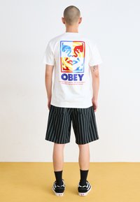 White t-shirt featuring a colorful graphic design with abstract faces and the word "OBEY," paired with black pinstripe shorts and black shoes.