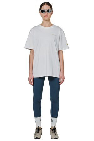 Kobieta w oversize białym T-shircie, ciemnoniebieskich legginsach, białych skarpetkach, beżowych sneakersach i dużych czarnych okularach przeciwsłonecznych, stojąca twarzą do przodu.