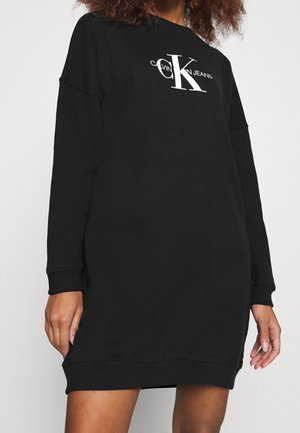 Kobieta ubrana w czarną sukienkę dresową Calvin Klein Jeans z długimi rękawami i białym napisem logo na piersi.