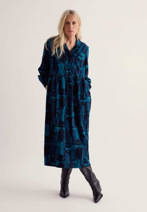 Femme blonde portant une longue robe bleue à motifs avec des boutons sur le devant et des bottes noires montant jusqu'aux genoux, debout les mains dans les poches.