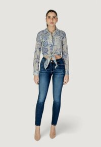 Lichtblauwe bloeiende cropped shirt met gestrikte voorkant, gecombineerd met donkere blauwe skinny jeans en nude hakken op een neutrale achtergrond.