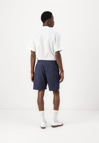 Hvit kortermet skjorte og marineblå shorts med elastisk midje, to baklommene, brukt med hvite joggesko og beige sokker.