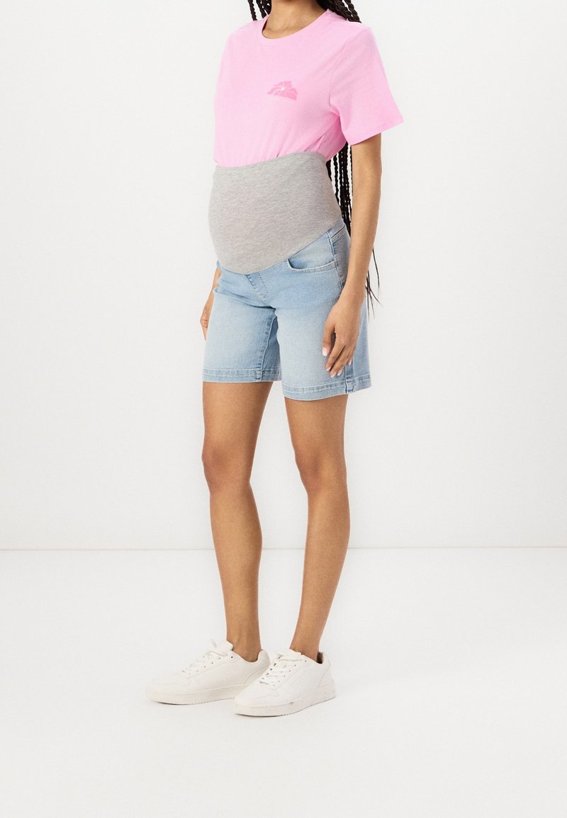 Růžové bavlněné tričko s logem, šedý pružný těhotenský pás, modré džínové kraťasy a bílé tenisky na neutrálním pozadí.