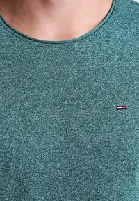 Green heathered t-shirt with a crew neckline. Features a small logo accent with red, white, and blue colors on the left chest area. Soft texture.