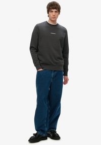 Grå sweatshirt med rund halsudskæring og broderet logo, parret med løse blå denimjeans og sorte snørebåndssko.