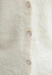 Jersey blanco esponjoso con una textura suave, que presenta cinco botones redondos con un acabado reflectante y bordes dorados.