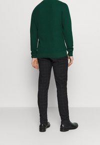 Homme portant un pull vert foncé, un pantalon slim noir à carreaux, et des bottes en cuir noir, debout face à un mur gris clair.