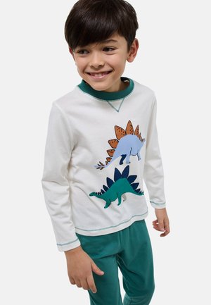 Garçon souriant portant une chemise blanche à manches longues avec deux dinosaures colorés et un pantalon vert, debout devant un fond clair uni.