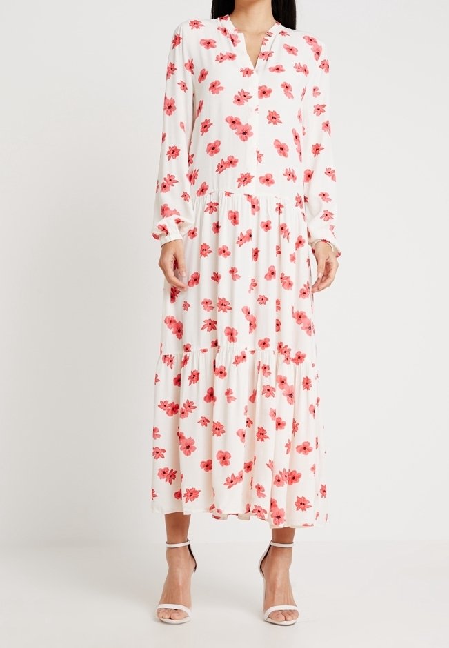 Femme portant une longue robe blanche avec des motifs floraux roses et des manches longues, assortie à des sandales blanches à talons hauts.