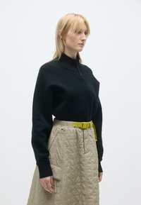 Melna trikotāžas džemperis ar augstu apkakli un rāvējslēdzēju, apvienots ar bēšas struktūras svārkiem, kuriem ir kabatas un spilgti dzeltena josta.