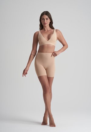 Vrouw staat blootvoets in beige naadloze onderkleding en wijst naar de high-waisted shorts tegen een neutrale achtergrond.