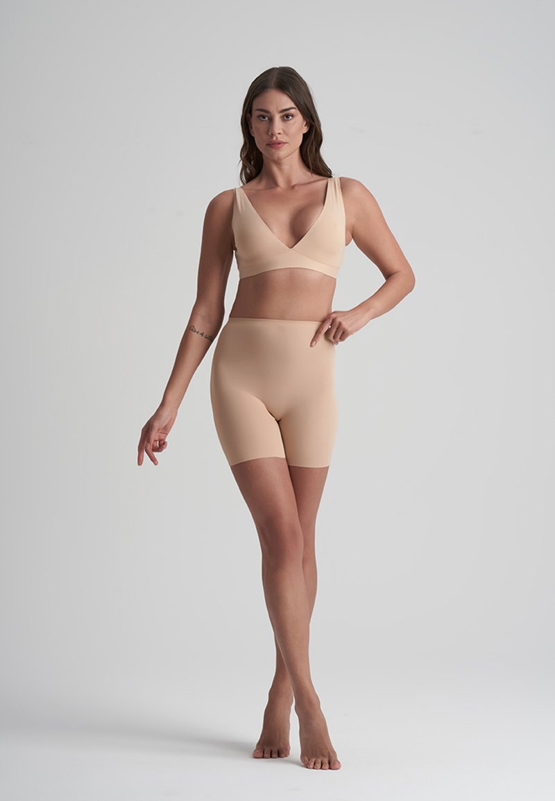 Kvinna som står barfota i beige sömlösa underkläder och pekar på de högmidjade shortsen mot en neutral bakgrund.