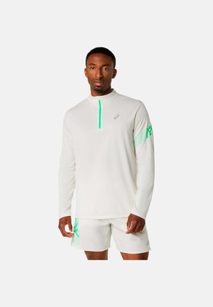 Uomo che indossa una maglia atletica bianca a maniche lunghe con cerniera e logo verdi, abbinata a pantaloncini coordinati con dettagli verdi.