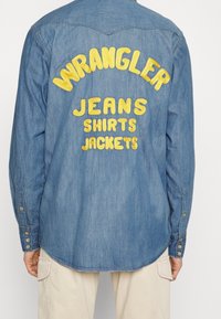 Camicia in denim con testo ricamato giallo sul retro: "WRANGLER JEANS SHIRTS JACKETS." Maniche lunghe con polsini a bottoni e orlo curvo.