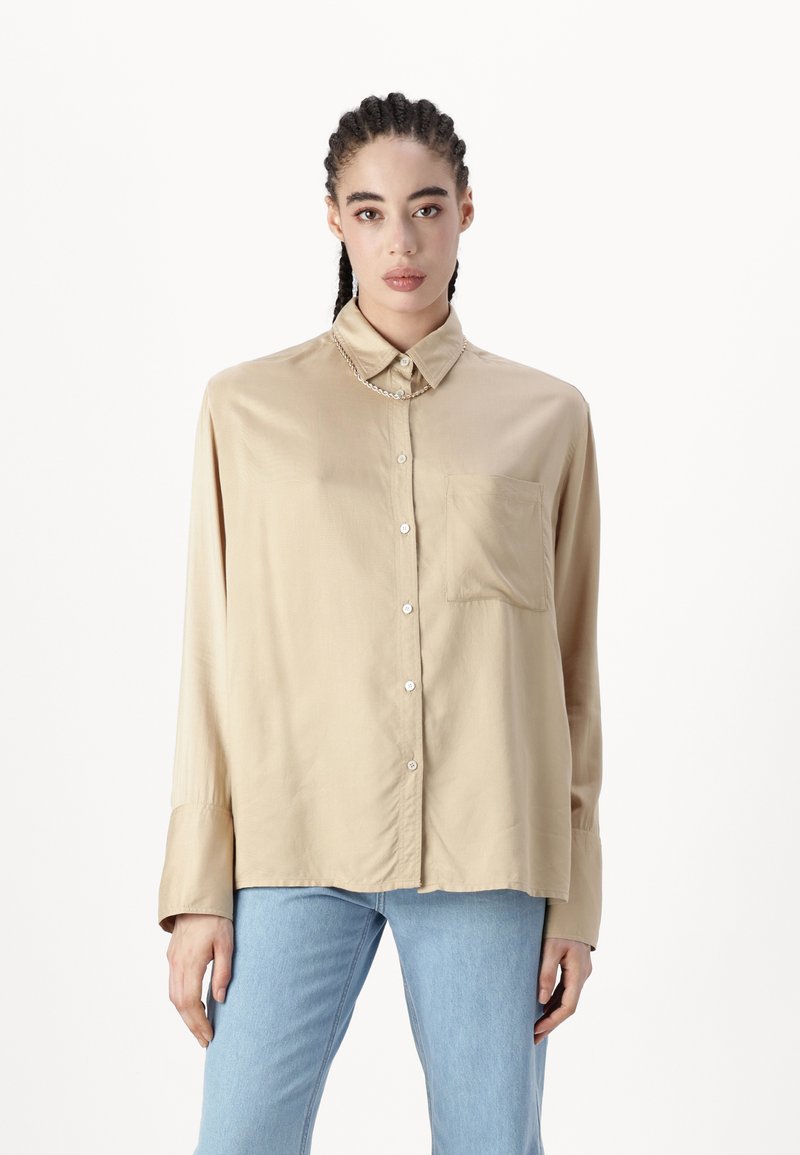 Beige långärmad skjorta i mjukt tyg, med krage, framknappar och bröstficka, i kombination med ljusblå jeans.