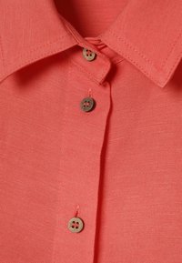 Col de chemise en lin rose corail avec trois boutons ronds en bois marron cousus avec un fil corail assorti.