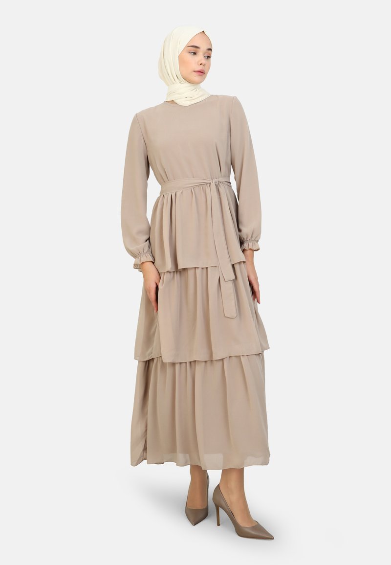 Beige, långärmad klänning med lager på lager, texturerat tyg, åtsittande midja och matchande bälte. Stylad med en ljusfärgad hijab och klackar.
