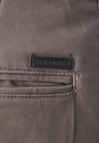 Tela marrón con una textura suave, que presenta un pequeño parche negro con el logo "JACK & JONES" encima de un bolsillo, acentuado por costuras visibles.
