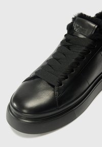 Baskets en cuir noir avec une semelle en caoutchouc texturé noir, de grosses lacets noirs et une doublure en fourrure douce autour de la cheville.