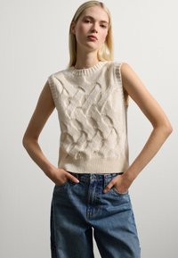 Ärmelloses Stricktop in Creme mit strukturiertem Zopfmuster, geripptem Rundhalsausschnitt und taillierter Passform. Kombiniert mit blauen, hoch geschnittenen Jeans.