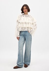 Blouse en dentelle blanche avec des volants en tiers, manches longues et un motif floral, associée à un jean large bleu clair et des chaussures marron.