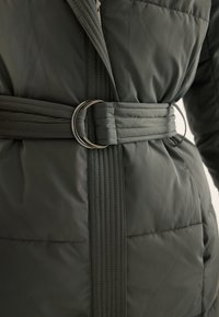 Szczegół szarej pikowanej kurtki z teksturowaną tkaniną, talią z paskiem zapinanym na srebrną okrągłą klamrę oraz przeszyciami.