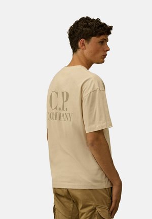 Giovane uomo con capelli corti e ricci, indossa una t-shirt beige C.P. Company e pantaloni cargo color sabbia, visto di profilo su uno sfondo semplice.