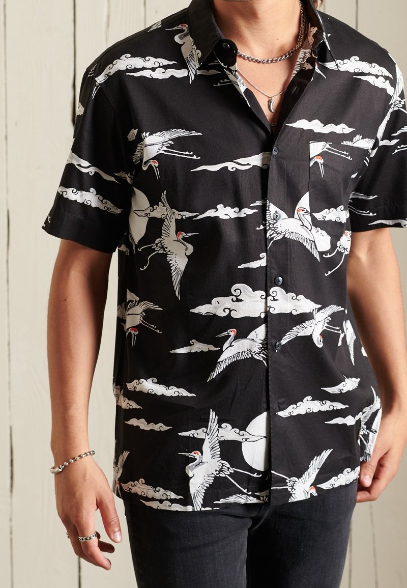 Chemise noire à manches courtes avec boutons, motifs de grues blanches et de nuages, portée par une personne avec un collier chaîne en argent et un bracelet.