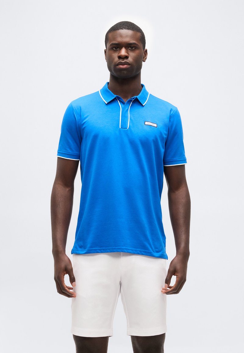 Homme debout de face portant un polo bleu vif avec des bordures blanches et un short blanc sur un fond uni.