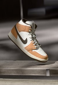 Beige, fehér és barna árnyalatú magas szárú sportcipő Nike logóval, fűzőkkel és perforált orral, levegőben, egy betonfelület felett.