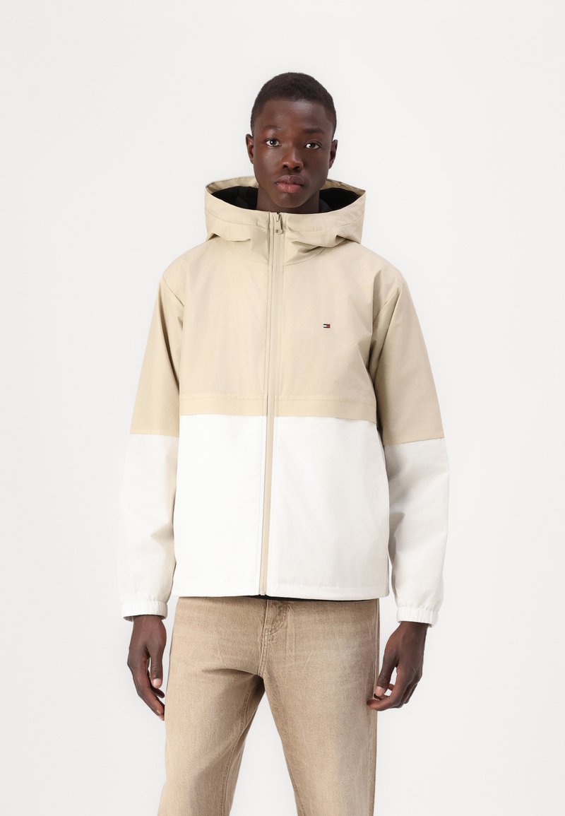 Beige and white hooded jacket with a zipper, featuring contrasting panels and a small logo on the chest. Smooth texture, lightweight fabric.